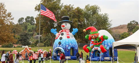 Snowman and elf bounce house next to American flag at a park with participants